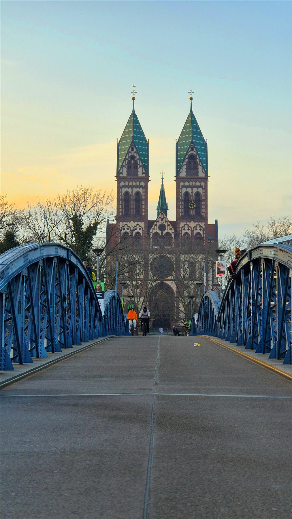 A blue steel bridge leading towards a twin-spired church